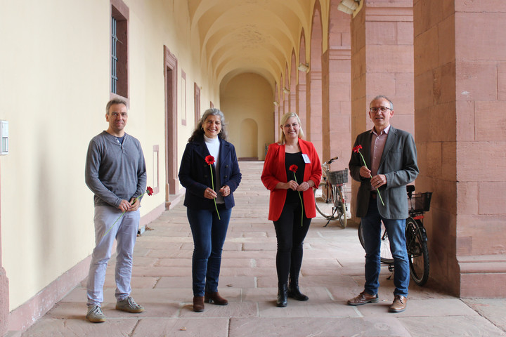 Vier Menschen stehen in einer Vorhalle des Mannheimer Schlosses. Alle lächeln und halten jeweils eine rote Blume.