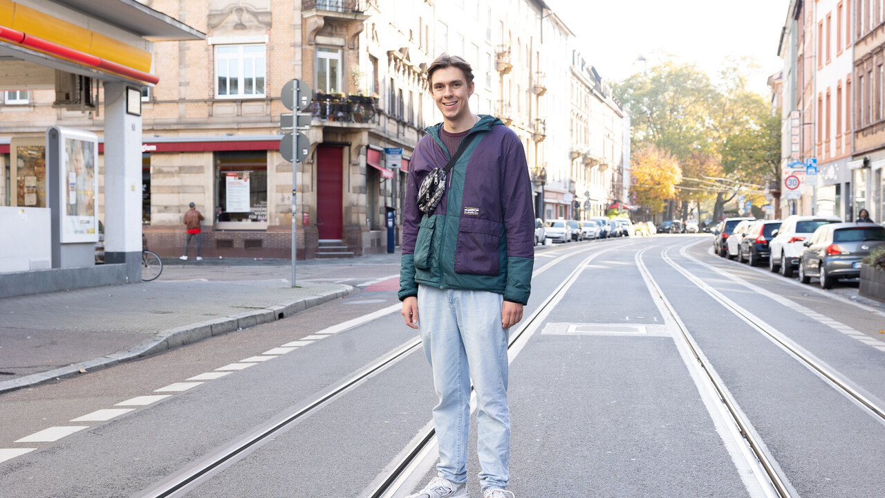 Ein Student mit einer lilanen sportlichen Jacke steht lachend auf Bahngleisen in der Schwetzinger Vorstadt.
