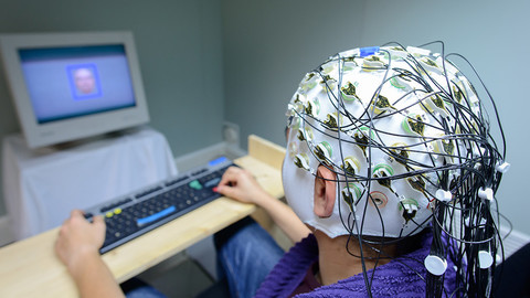 Eine Person trägt eine Haube mit Elektroden auf dem Kopf und sitzt an einem Schhreibtisch. Sie blickt in einen Bildschirm und tippt auf einer Tastatur.