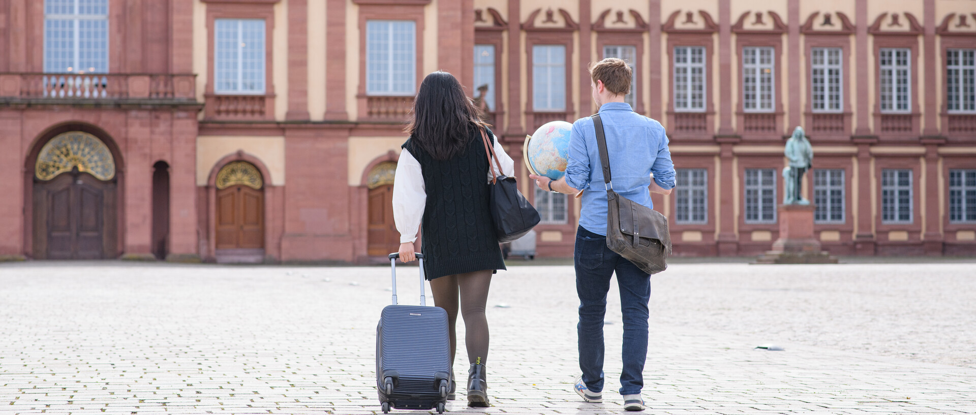 Zwei Personen laufen nebeneinander auf das Schloss der Uni Mannheim zu. Eine Person zieht einen schwarzen Koffer, während die andere Person eine Tasche sowien einen Globus trägt.