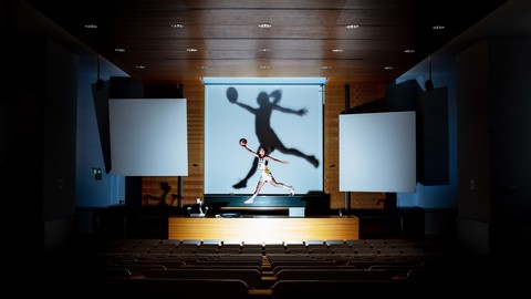Eine Person trägt Sportkleidung und springt über einem Pult in einem leeren Hörsaal. In ihren rechten Hand hält sie ausgestreckt einen Handball.