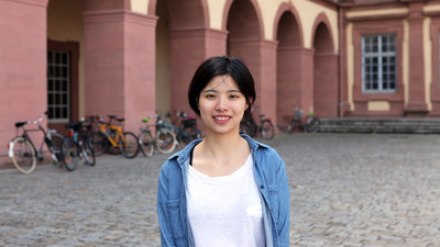 Eine lächelnde Person trägt ein weißes T-Shirt sowe ein blaues Jeanshemd und steht auf dem Ehrenhof. Die Person heißt Jianing Shen.