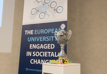 Ein silberner Pokal steht auf einem Regal. Hinter dem Regal steht ein Banner, auf dem folgendes zu lesen ist: "The european university engaged in societal changes".