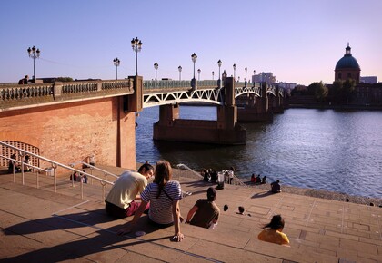 Einige Personen sitzen auf Stufen neben einem Fluss. Links von den Stufen ist eine Brücke.