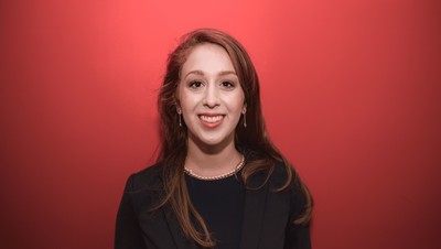 Eine lächelnde Person trägt ein schwarzes Top sowie einen schwarzen Blazer und steht vor einem roten Hintergrund. Die Person heißt Maya Moritz.