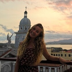 Djamila Klöfer trägt ihre langen lockigen blonden Haare offen. Im Hintergrund ein Sonnenuntergang und der Blick auf Havanna.