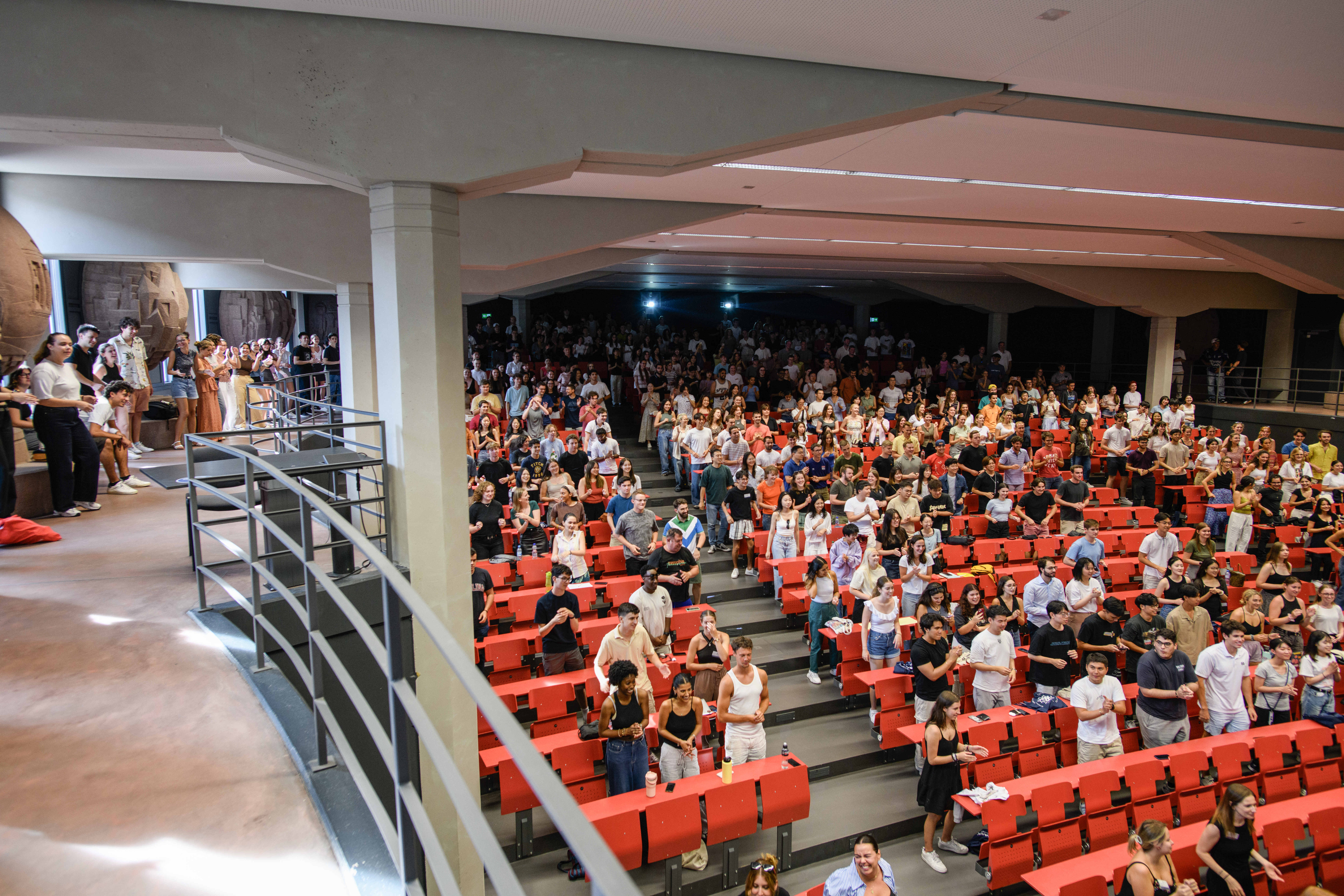 Many students are standing in a lecture hall with red furniture.