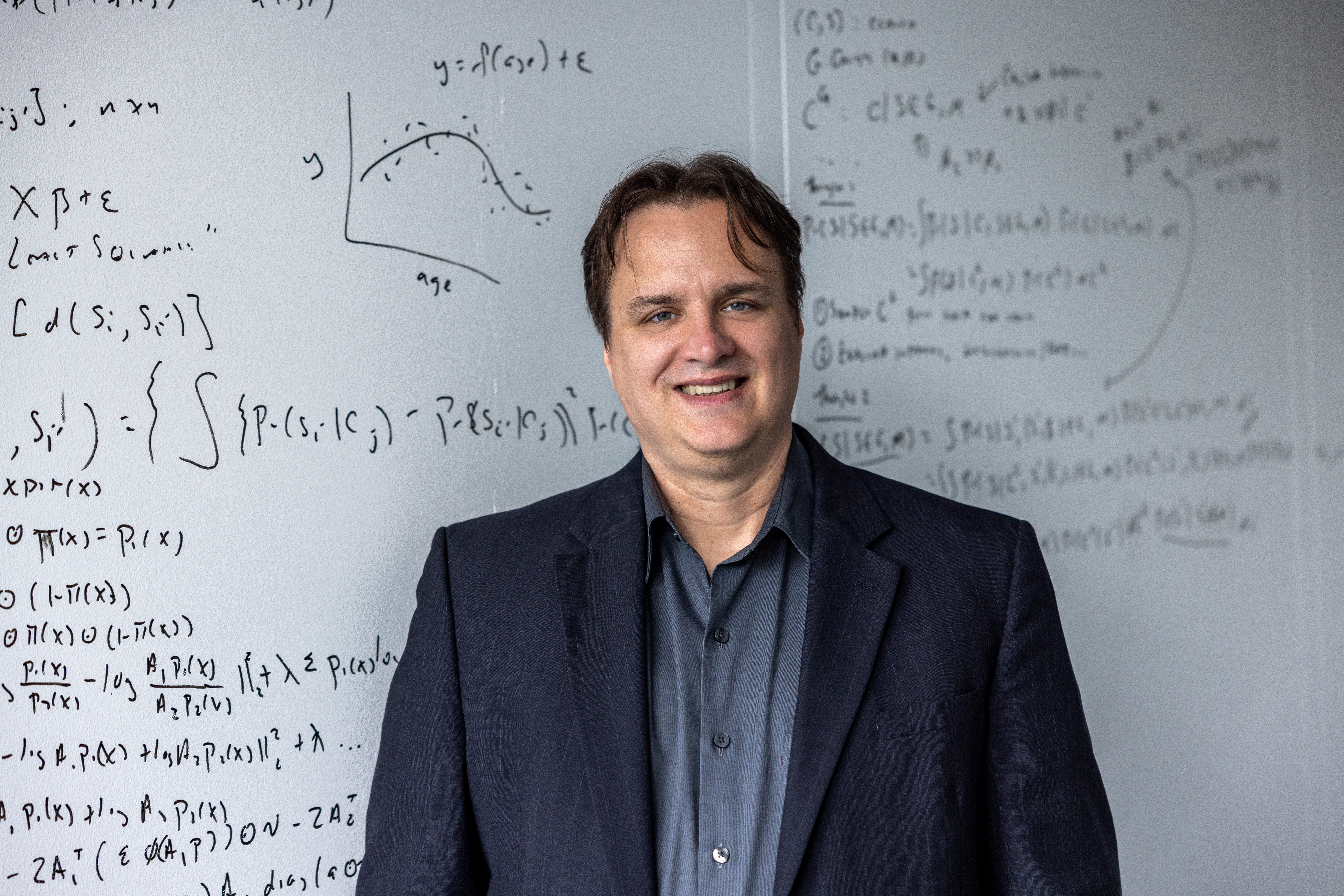 Portrait von Marc Ratkovic. Er trägt ein graues Hemd sowie einen dunkleblauen Blazer und steht lächelnd vor einem Whiteboard.