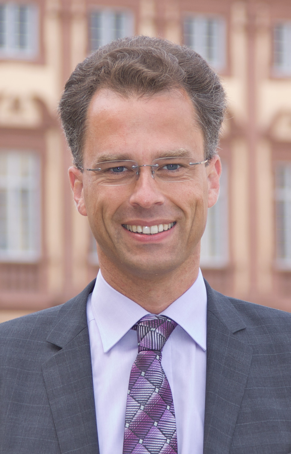 Portrait von Georg Bitter. Er tträgt ein weißes Hemd, eine lila-farbene Krawatte sowie einen grauen Blazer und steht vor dem Schloss.