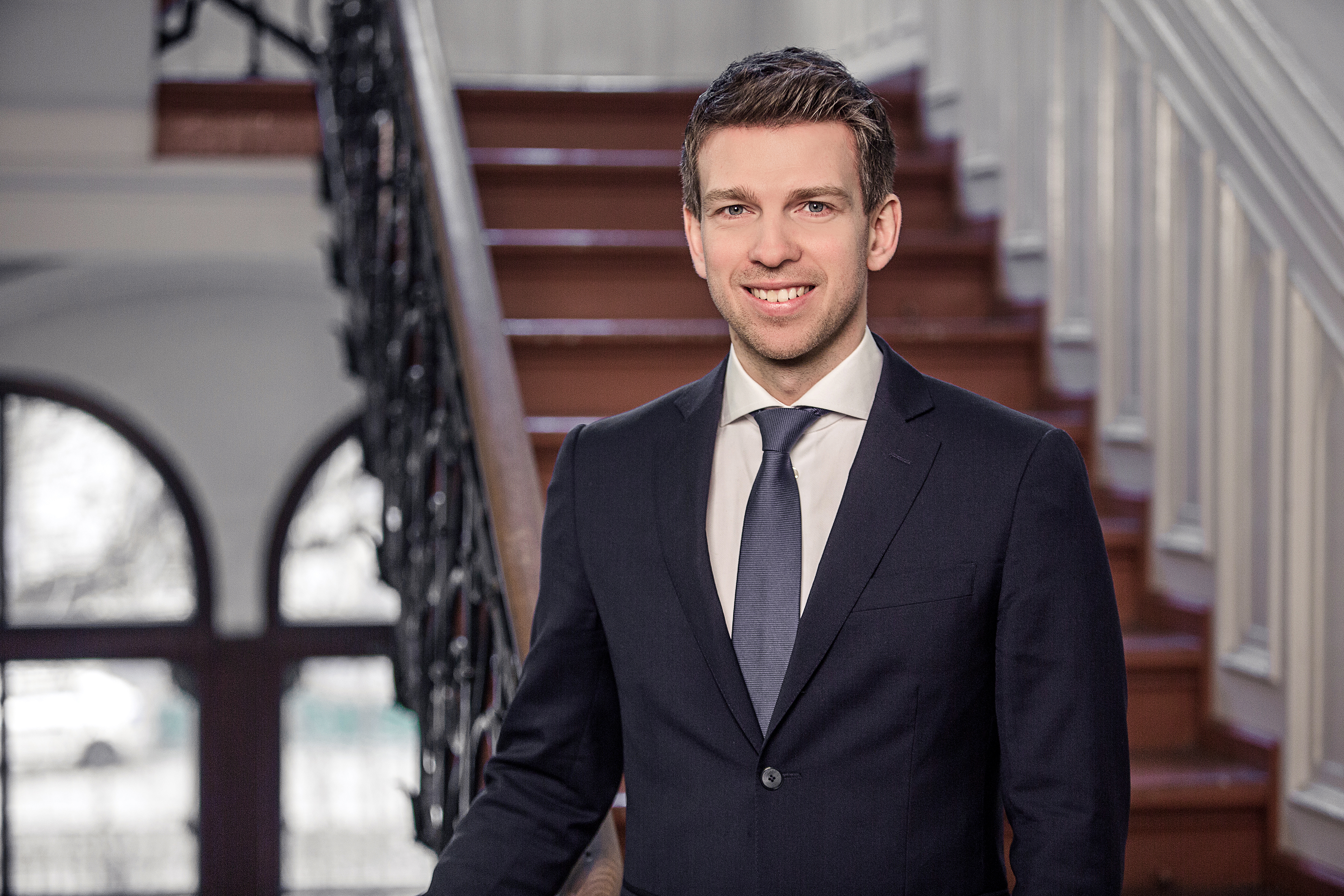 Portrait von Arnd Vomberg. Er trägt ein weißes Hemd, eine dunkelblaue Krawatte sowie einen dunkelblauen Blazer. Er steht in einem Treppenhaus.