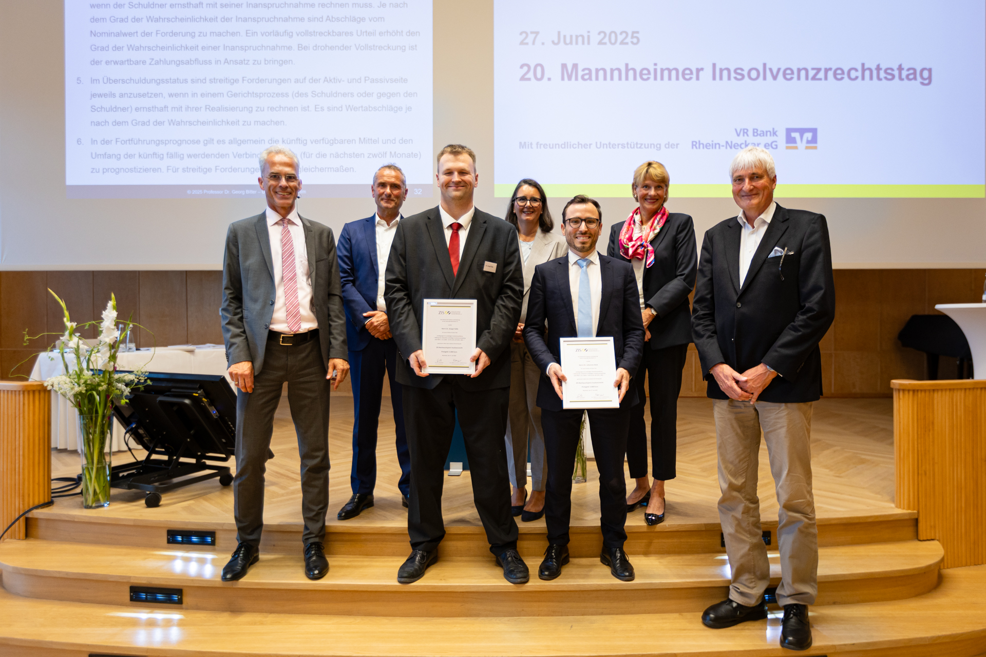 Gruppenbild mit den Preisträgern Dr. Johannes Heck und Dr. Ansgar Kalle mit den Mitglieder der Auswahlkommission Prof. Dr. Georg Bitter, RA Tobias Hoefer, Vors. RiBGH a.D. Prof. Dr. Godehard Kayser, RAin Annette Kollmar, RAin Dr. Alexandra Schluck-Amend. Alle Personen sind mit Anzügen gekleidet und stehn auf einer Bühne. Die beiden Preisträger zeigen ihre Urkunden.
