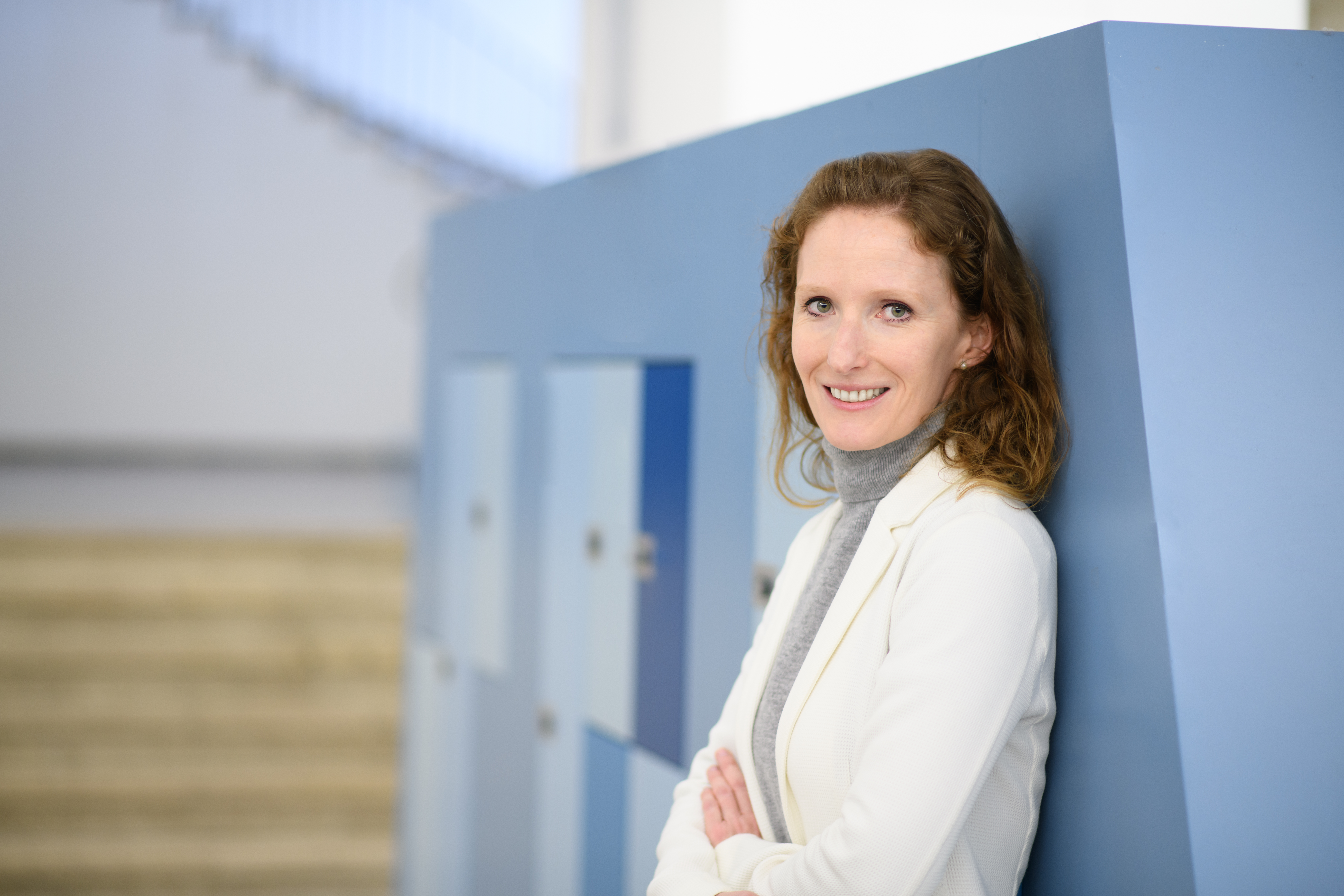 Porträt von Karin Hoisl. Sie hat braune, halblange Haare, trägt ein weißes Jacket und lächelt in die Kamera. Sie lehnt an einer Wand mit blauen Schließfächern.