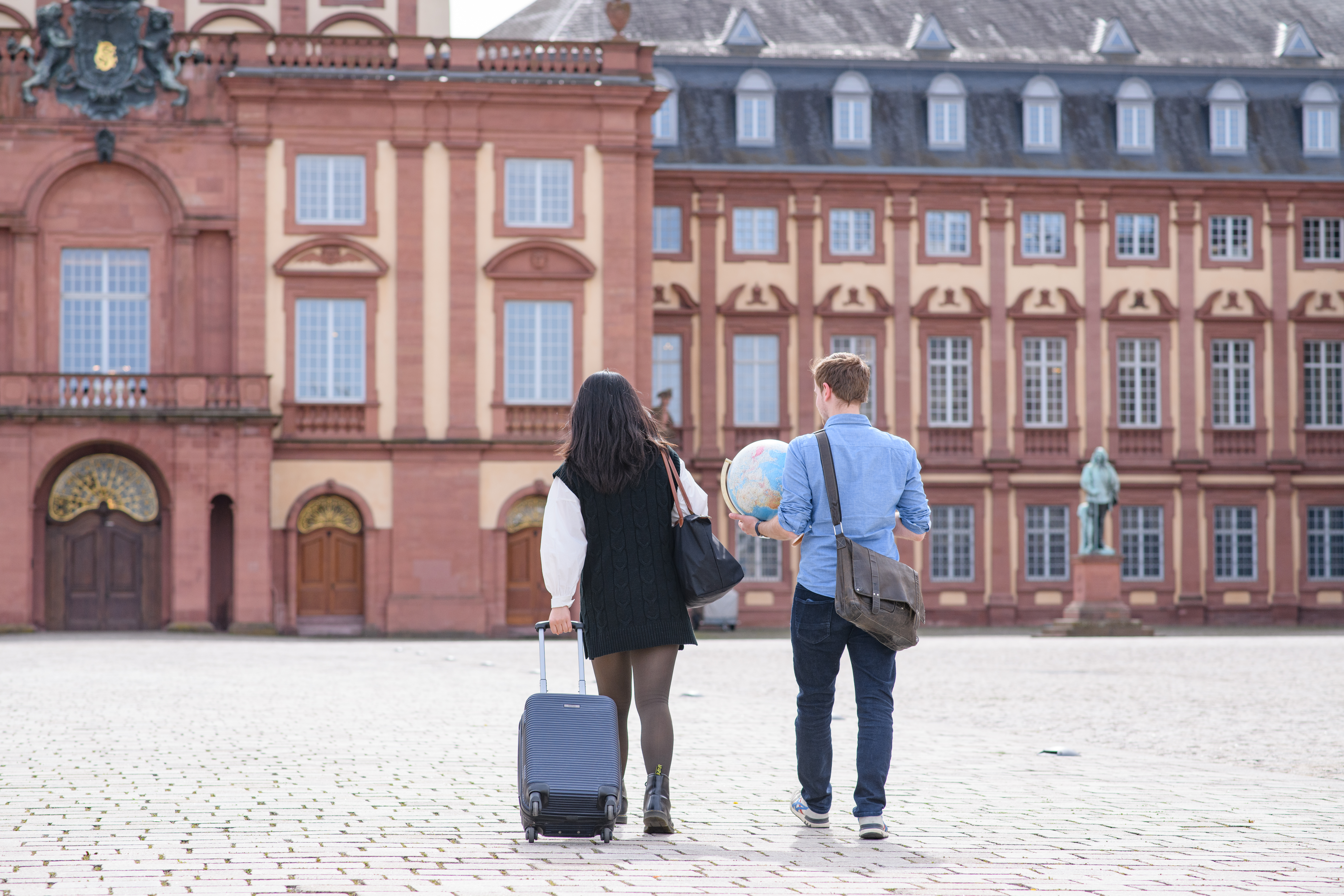 Two people walk side by side towards the Schloss of the University of Mannheim. One person pulls a black suitcase while the other person carries a bag and a globe.
