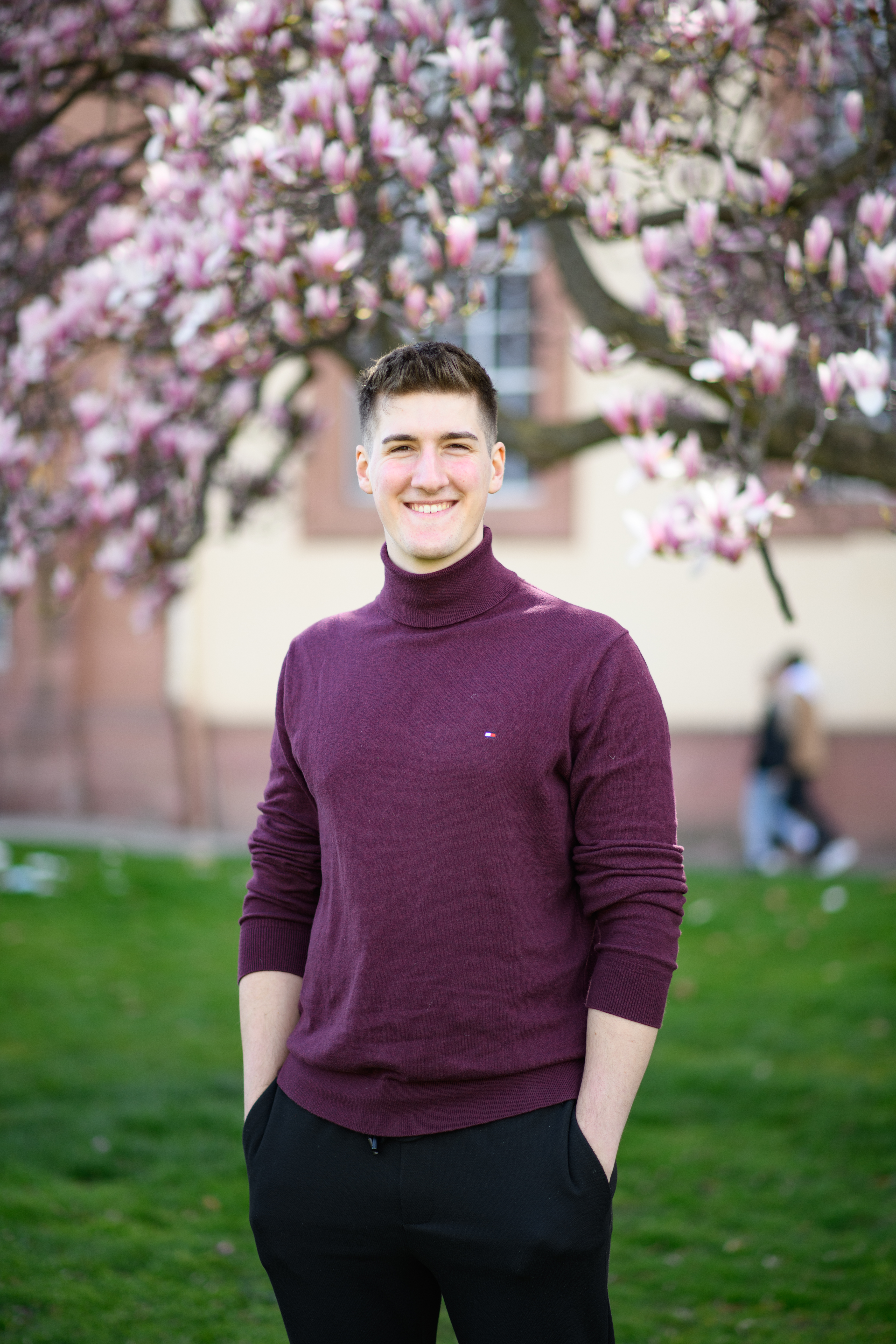 Eine lächelnde Person trägt einen weinroten Pullover sowie eine schwarze Hose und steht auf einer Wiese vor einem Magnolienbaum. Die Person heißt Jonas Wenzel.