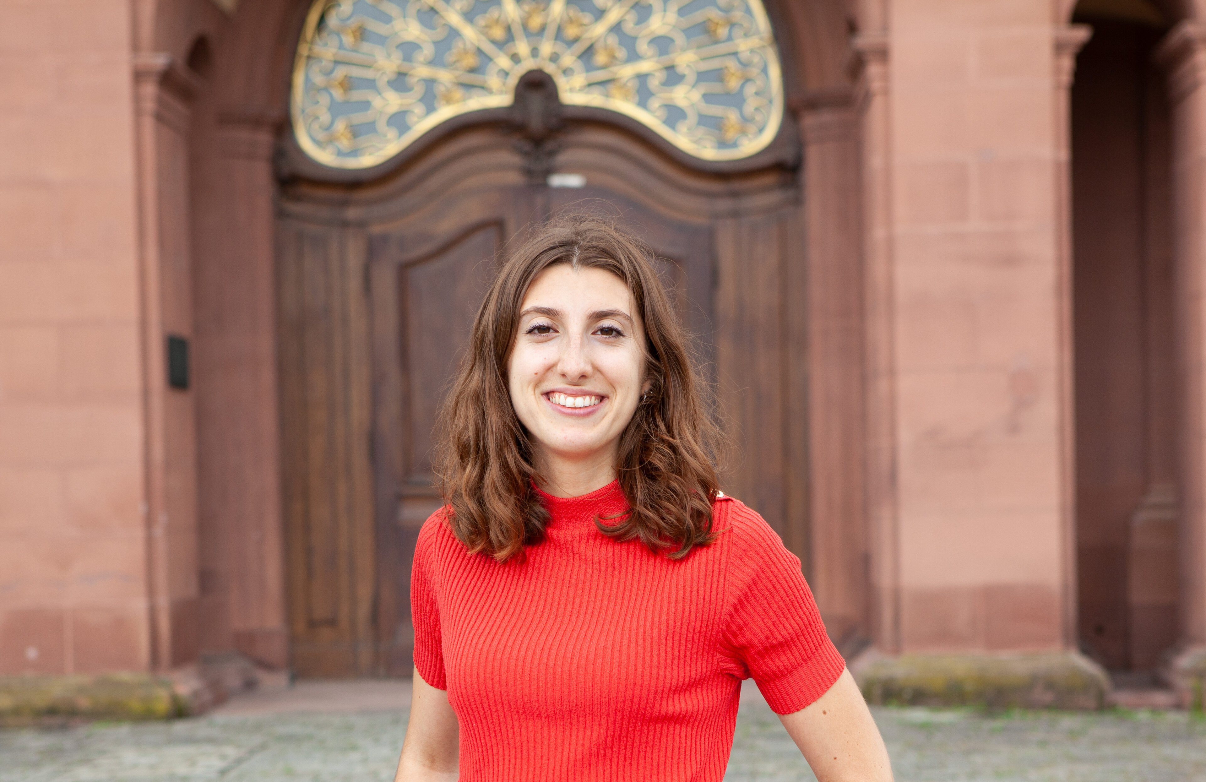 Eine lächelnde Person trägt ein rotes T-Shirt und steht vor einer braunen Schlosstüre. Die Person heißt Léa Beringuer.
