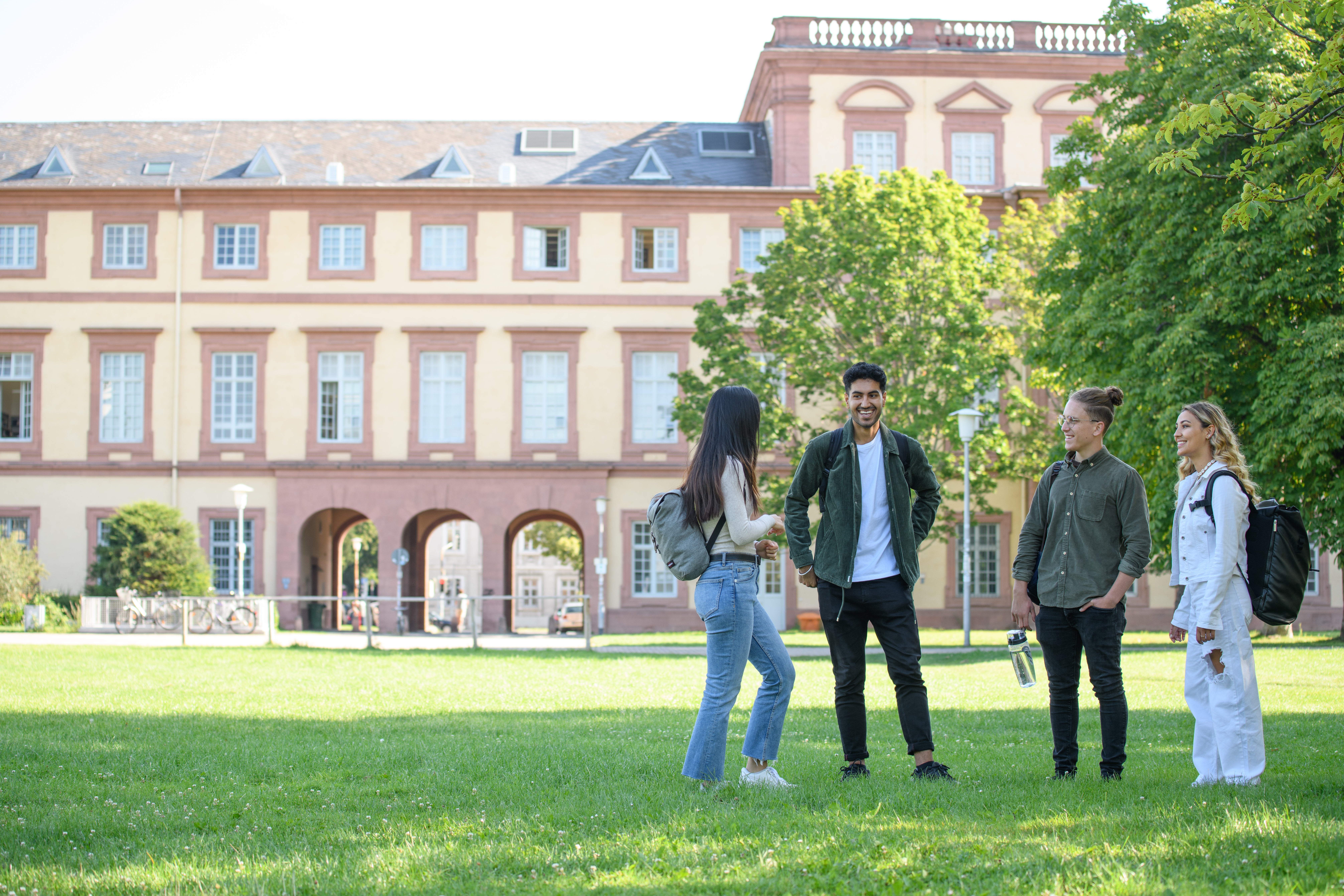 Vier Studierende stehen auf der Mensawiese. Die Studentin links hat lange braune Haare und trägt ein weißes Oberteil. Der Student rechts daneben hat kurze braune Haare und trägt ein weißes Oberteil mit einer dunklen Jacke. Der Student rechts neben ihm hat seine dunklen Haare zu einem Zopf gebunden und trägt ein grünes Oberteil. Die Studentin rechts neben ihm hat lange blonde Haare und trägt eine weiße Jacke.