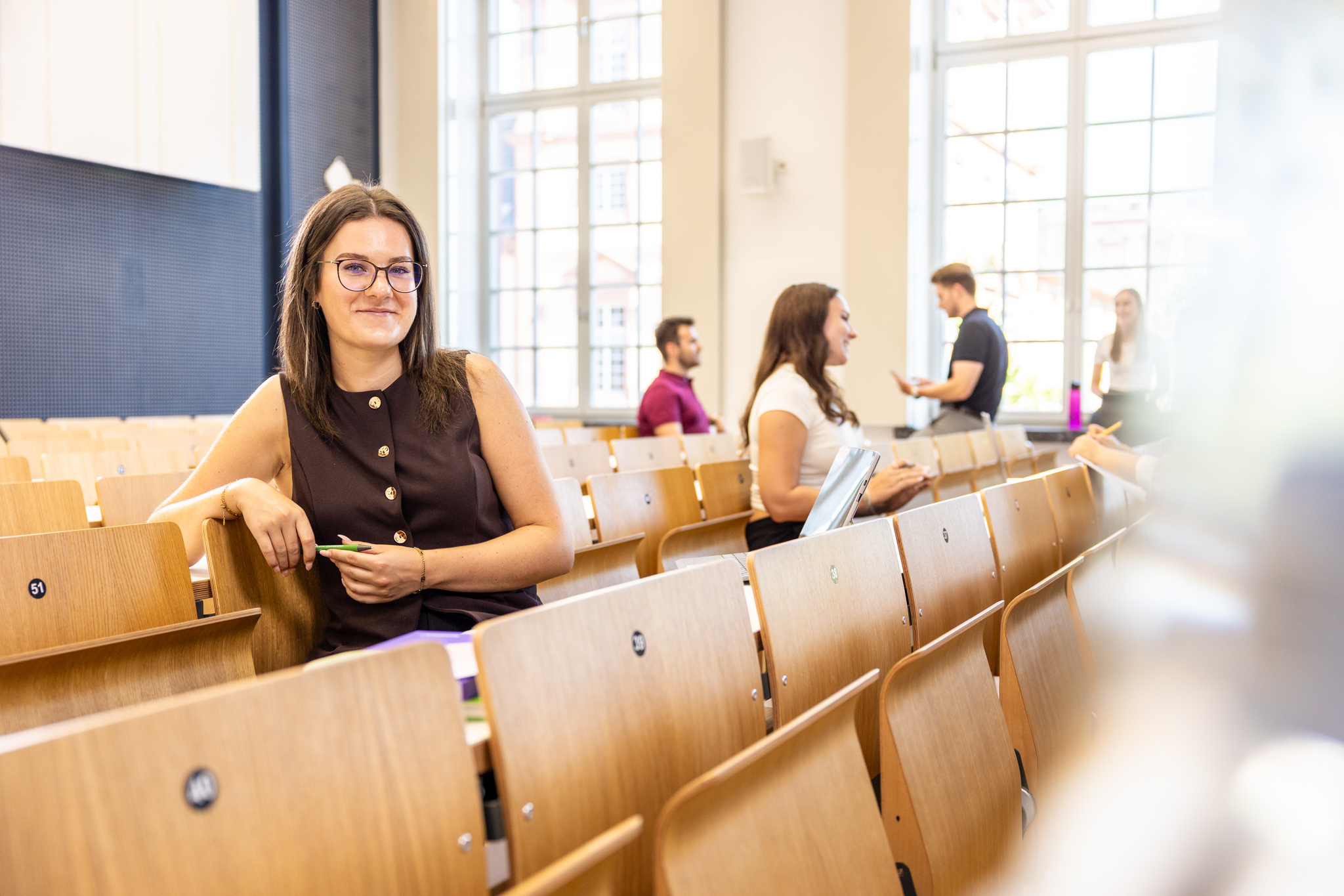 Eine Studentin sitzt in einer Bankreihe im Hörsaal. Sie schaut direkt in die Kamera und lächelt. Im Hintergrund sitzen weitere Studierende.