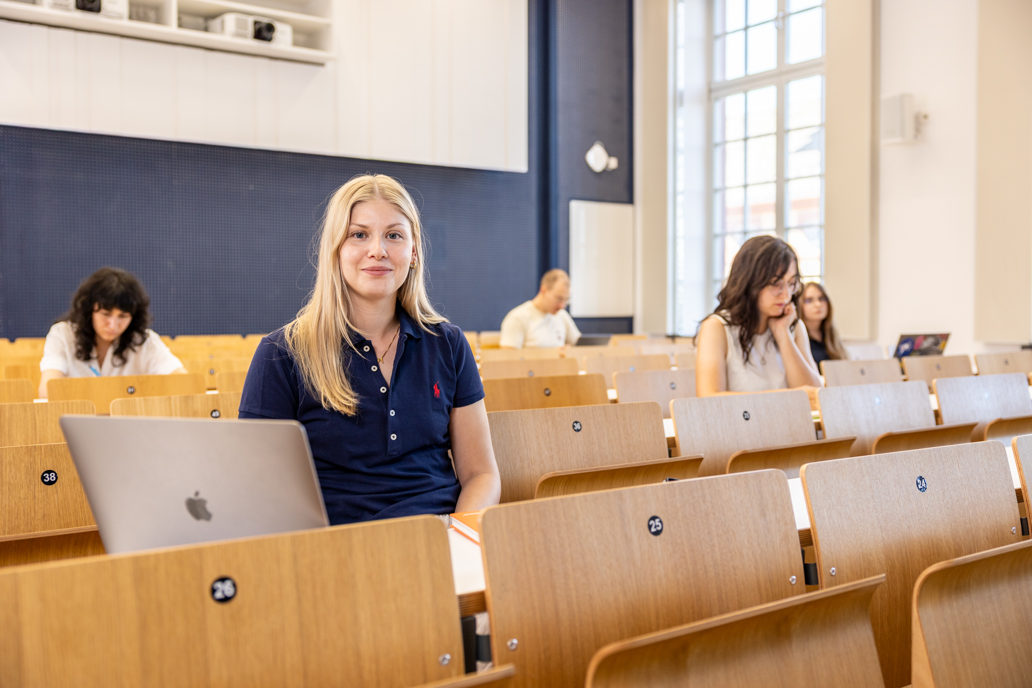 Eine Studentin sitzt in einer Bankreihe im Hörsaal. Vor sich hat sie ihren aufgeklappten Laptop. Sie schaut direkt in die Kamera und lächelt. Im Hintergrund sitzen weitere Studierende.
