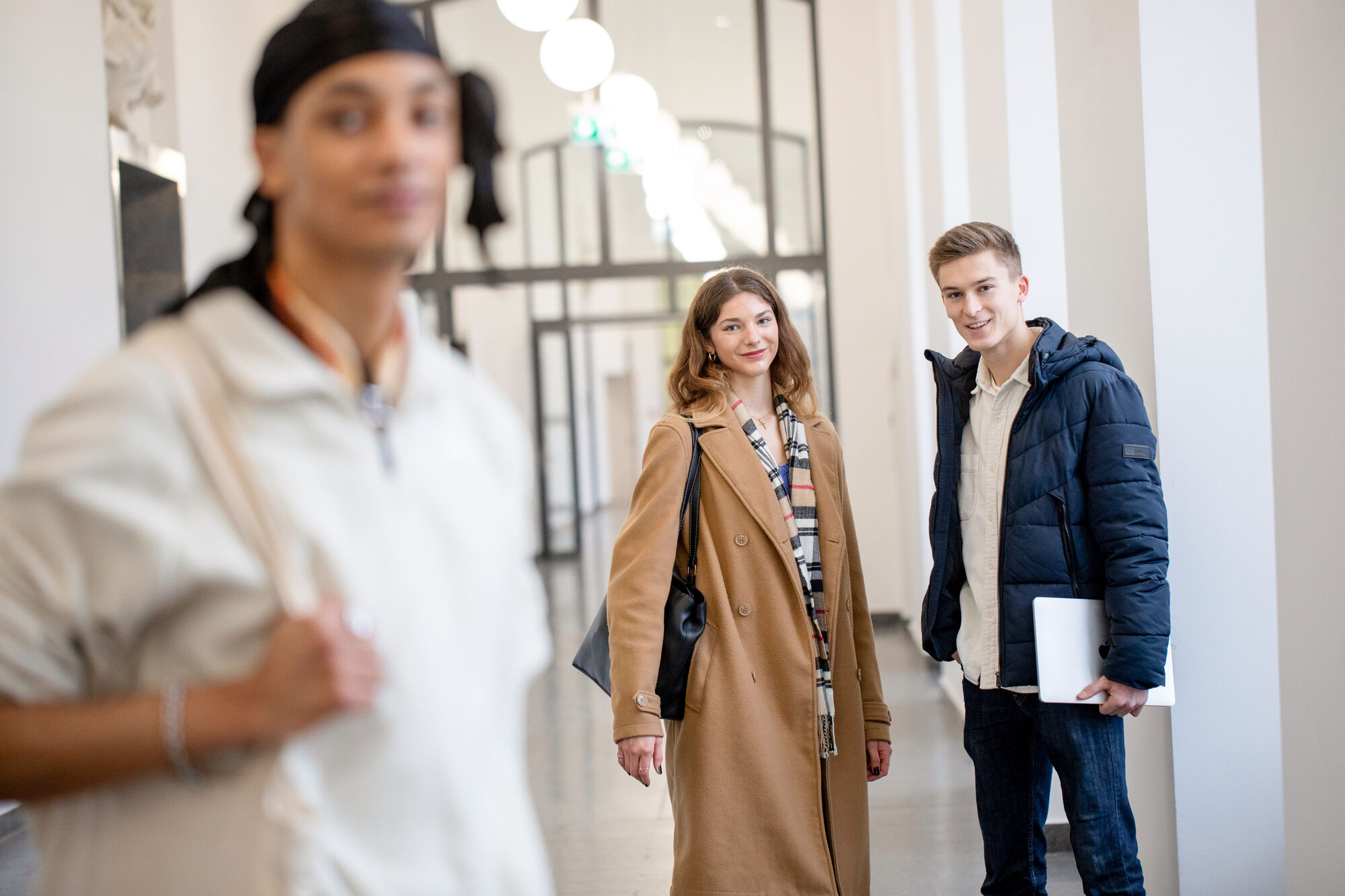 Eine Studentin und ein Student stehen im Flur des Schlosses. Sie tragen Jacken und Laptops unter dem Arm. Sie schauen interessiert in die Kamera.