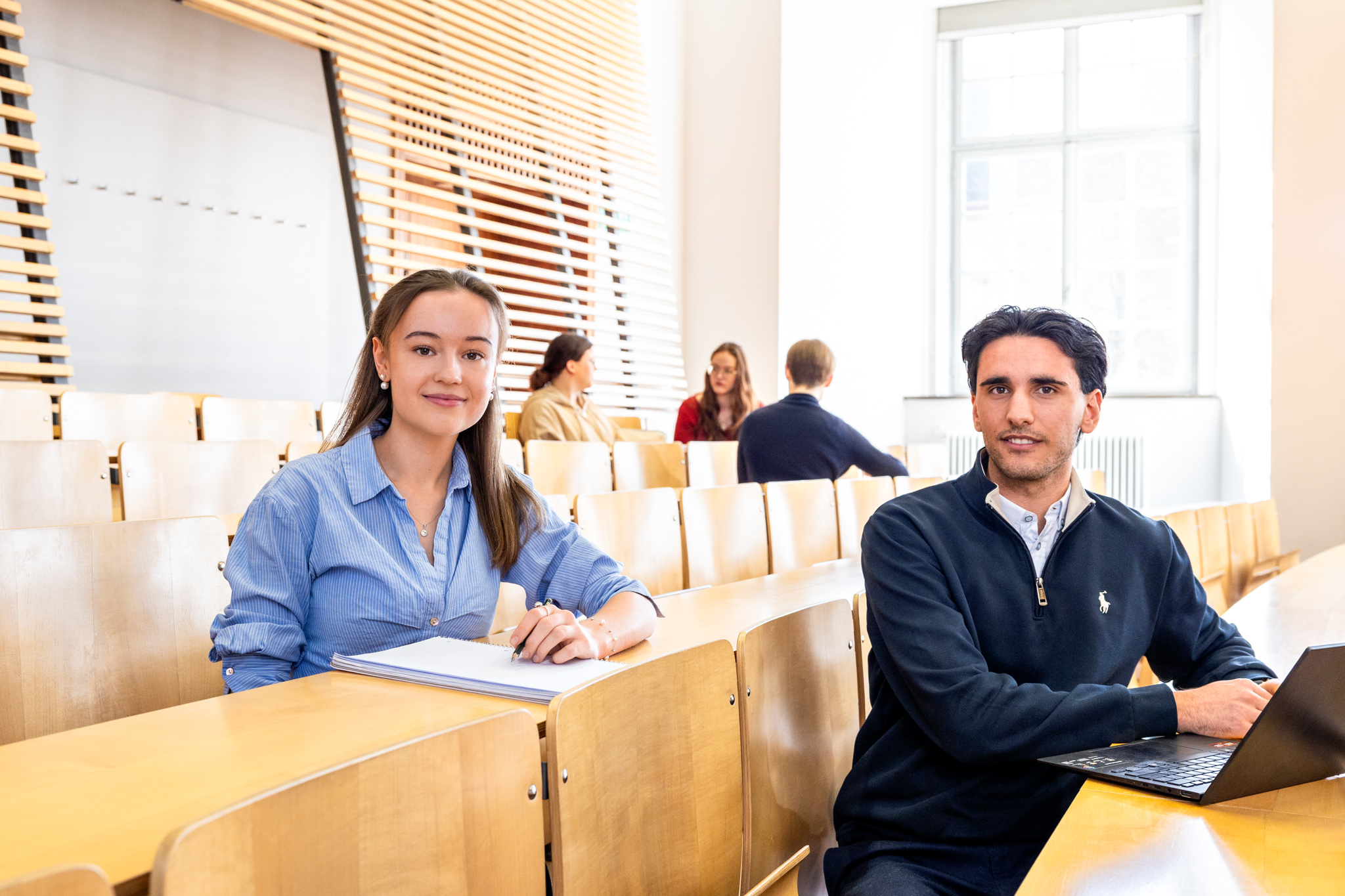 Studierende sitzen in einem Hörsaal. Sie schauen direkt in die Kamera.