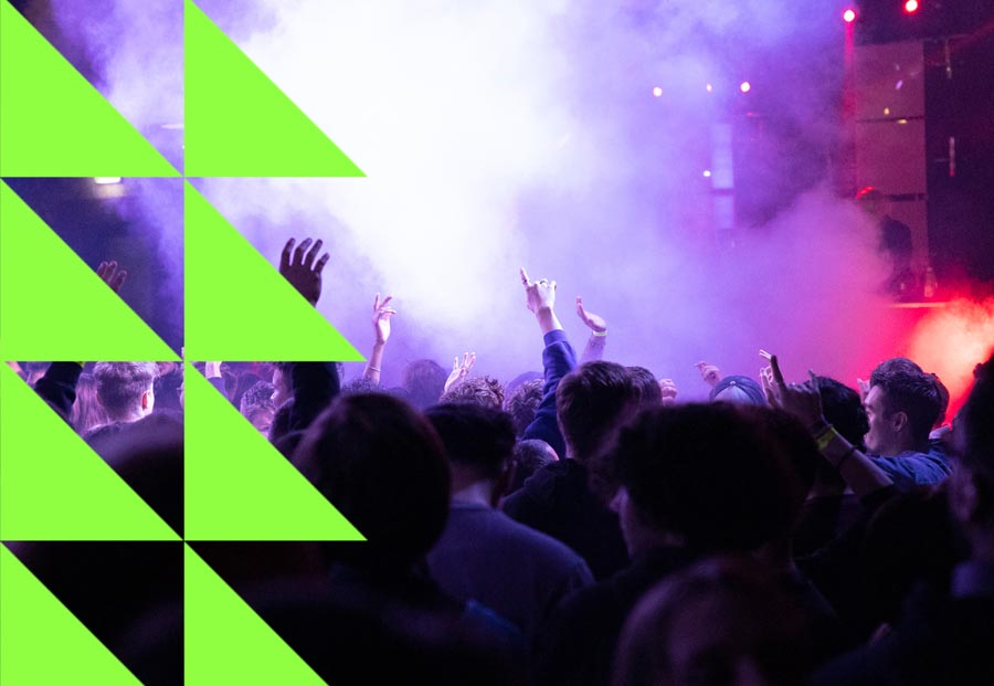 A crowd is raising their hands at a party, it is dark and colorful lights are illuminating the scene.