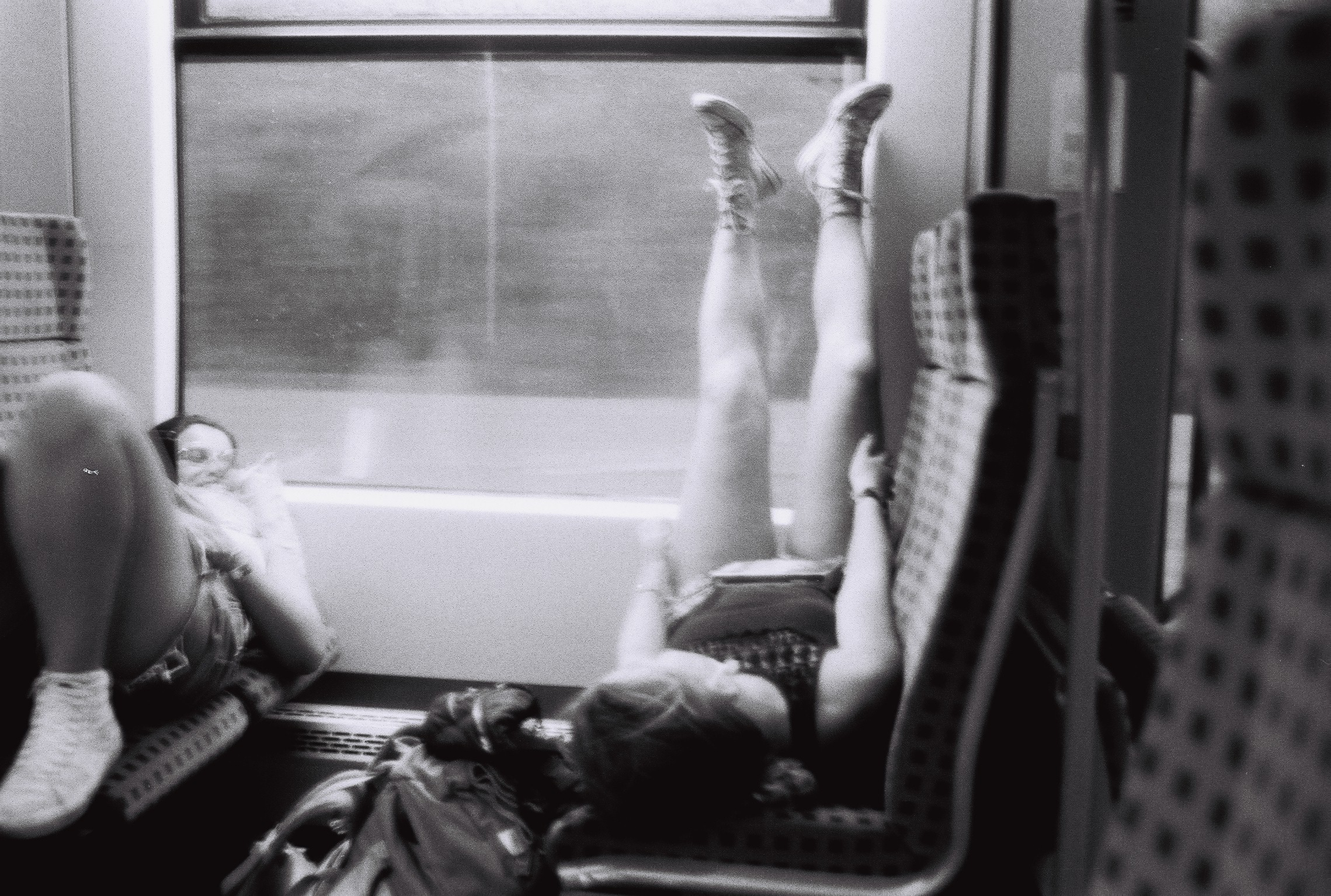 Zwei Frauen liegen auf Sitzen in einem Zug, eine der Frauen hat ihre Beine gegen die Schreibe gedrückt. Das Foto ist in Schwarz Weiß.