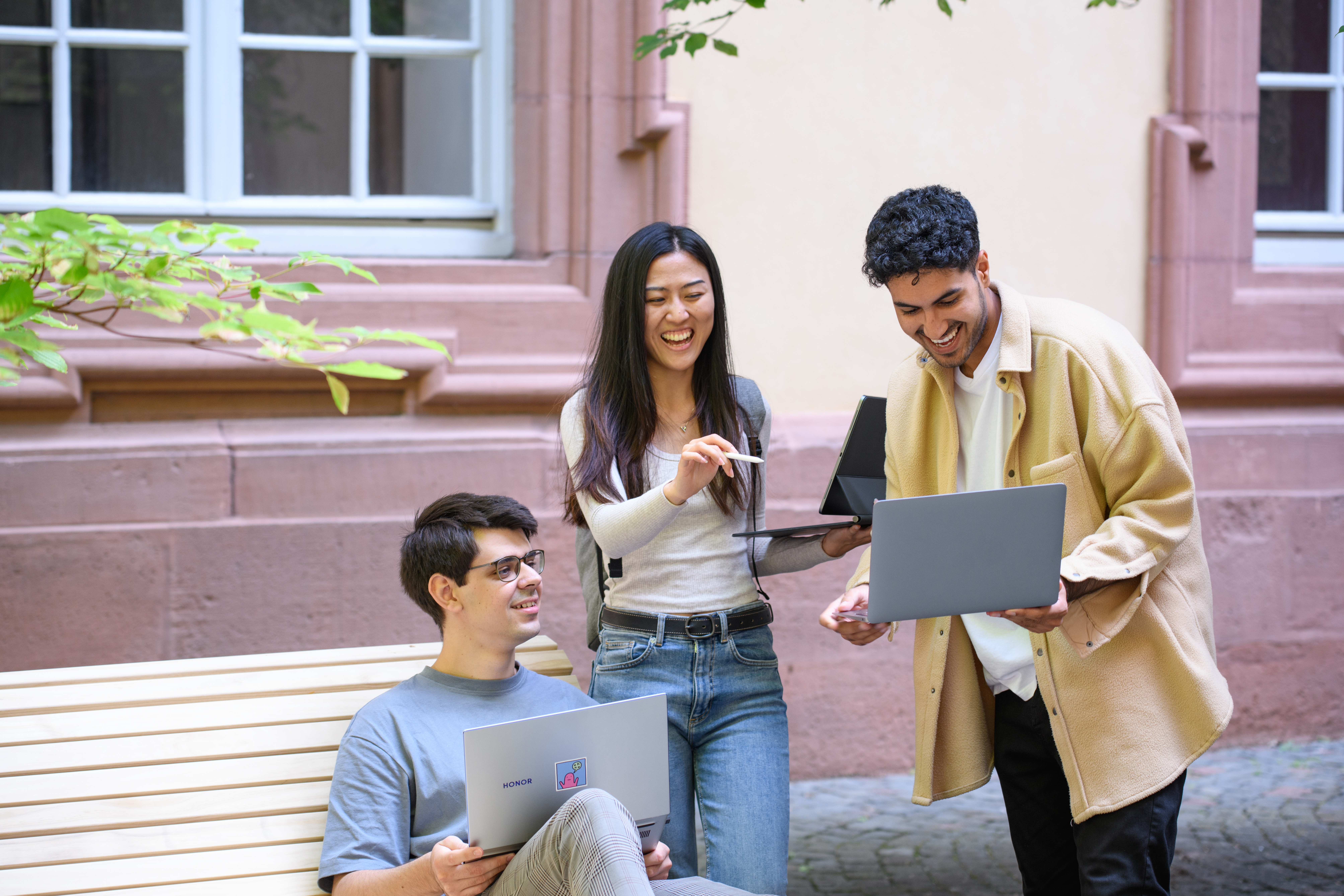 Drei Studierende halten Laptops in ihren Händen und lachen. 