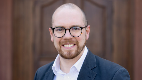 Benjamin Tödtmann. He is wearing a black suit and is standing in front of a brown door.