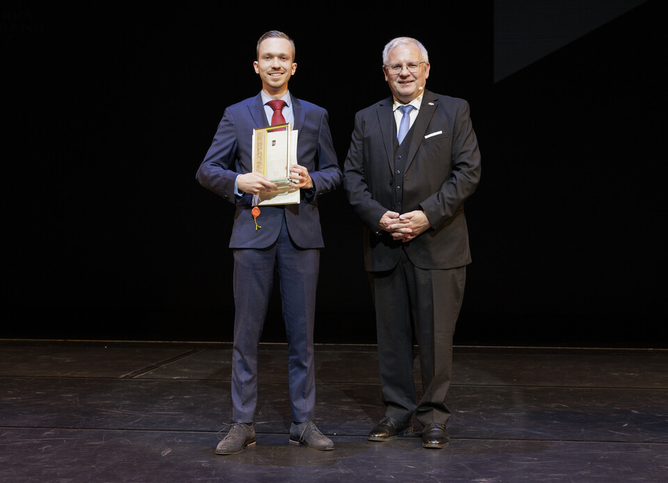 Auf einer Bühne vor schwarzem Hintergrund stehen auf der linken Seite Max Spanier mit dem Pfalzpreis und rechts der Bezirkstagsvorsitzende Hans-Ulrich Ihlenfeld. 