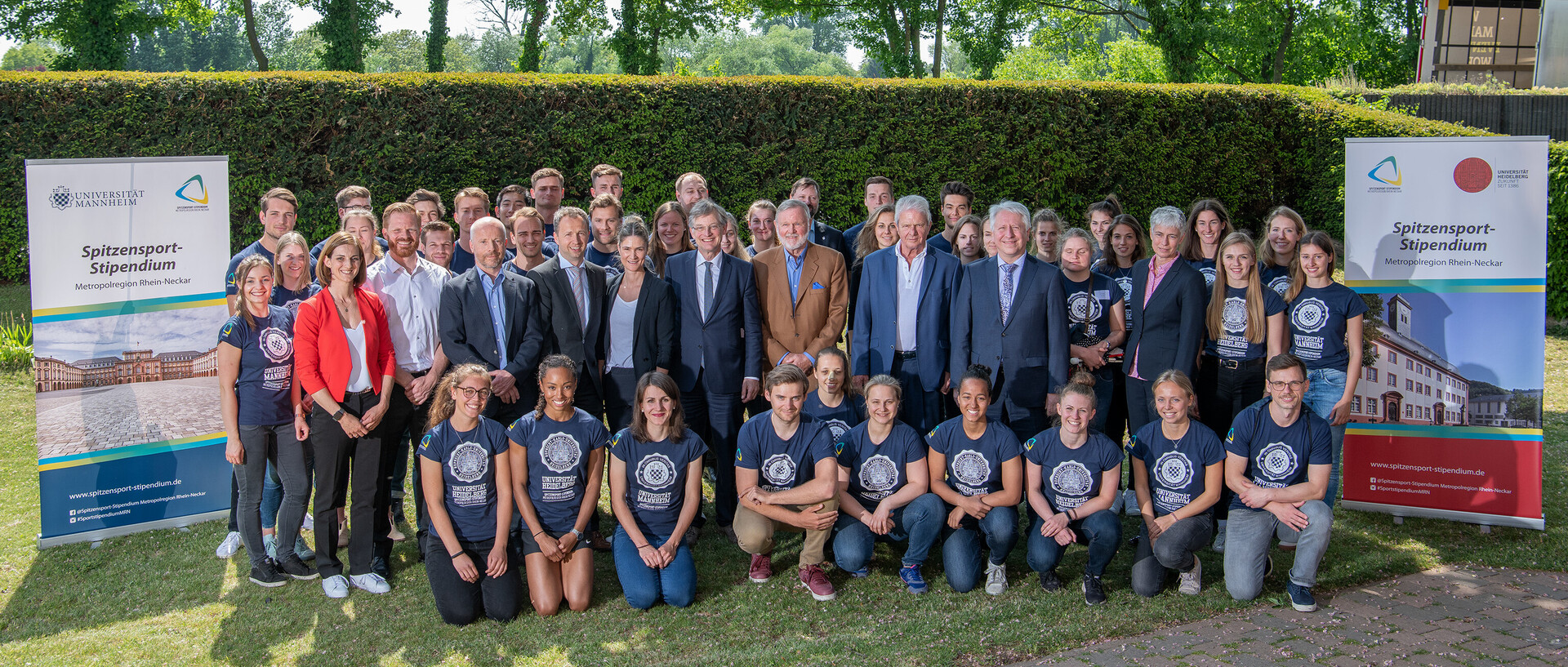 Eine Gruppe von Menschen steht auf einer Grünfläche. Rechts und links von der Gruppe steht ein Banner mit der Aufschrift "Spitzensport-Stipendium".