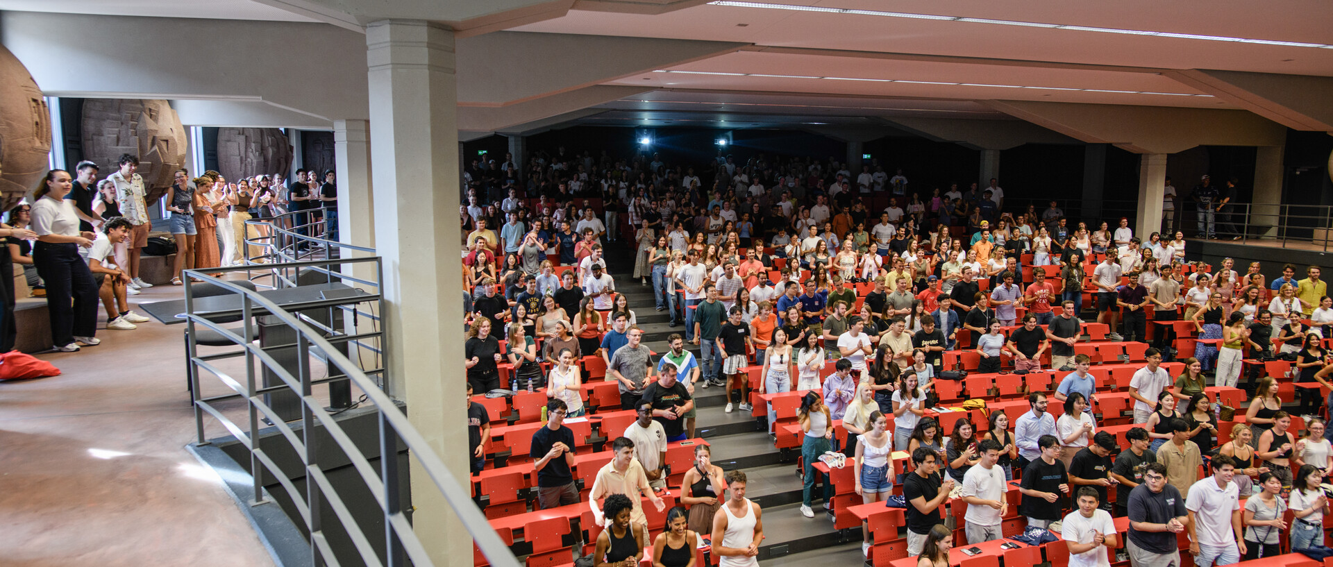 Viele Studierende stehen in einem Hörsaal mit roten Möbeln.