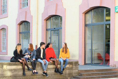 Fünf Studierende sitzen im Schneckenhof auf einer Mauer vor der Bibliothek.