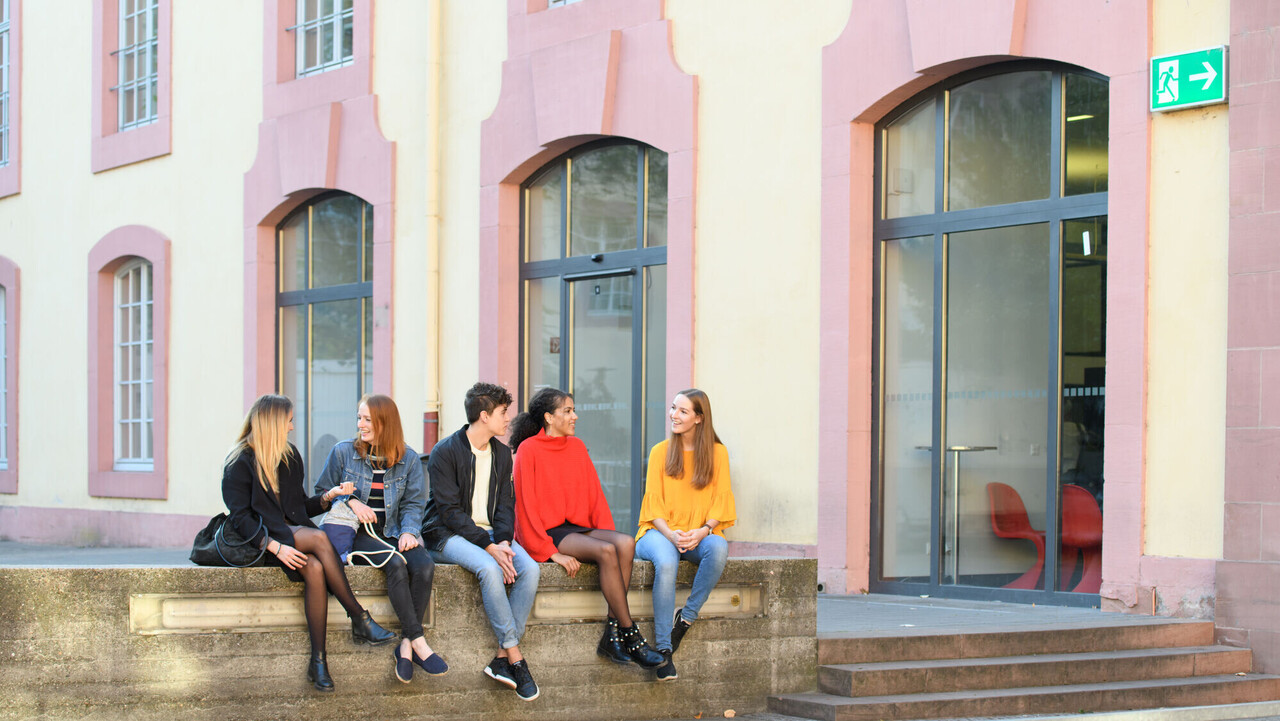 Fünf Studierende sitzen im Schneckenhof auf einer Mauer vor der Bibliothek.