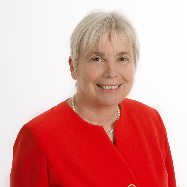 Angelika Storrer in roter Bluse und Perlenkette vor einem weißen Hintergrund.