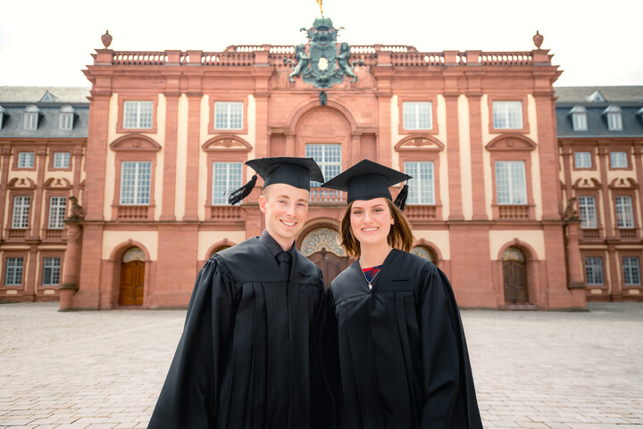 Zwei Studierende mit schwarzer Robe und Doktorhut stehen vor dem Schloss der Universität Mannheim
