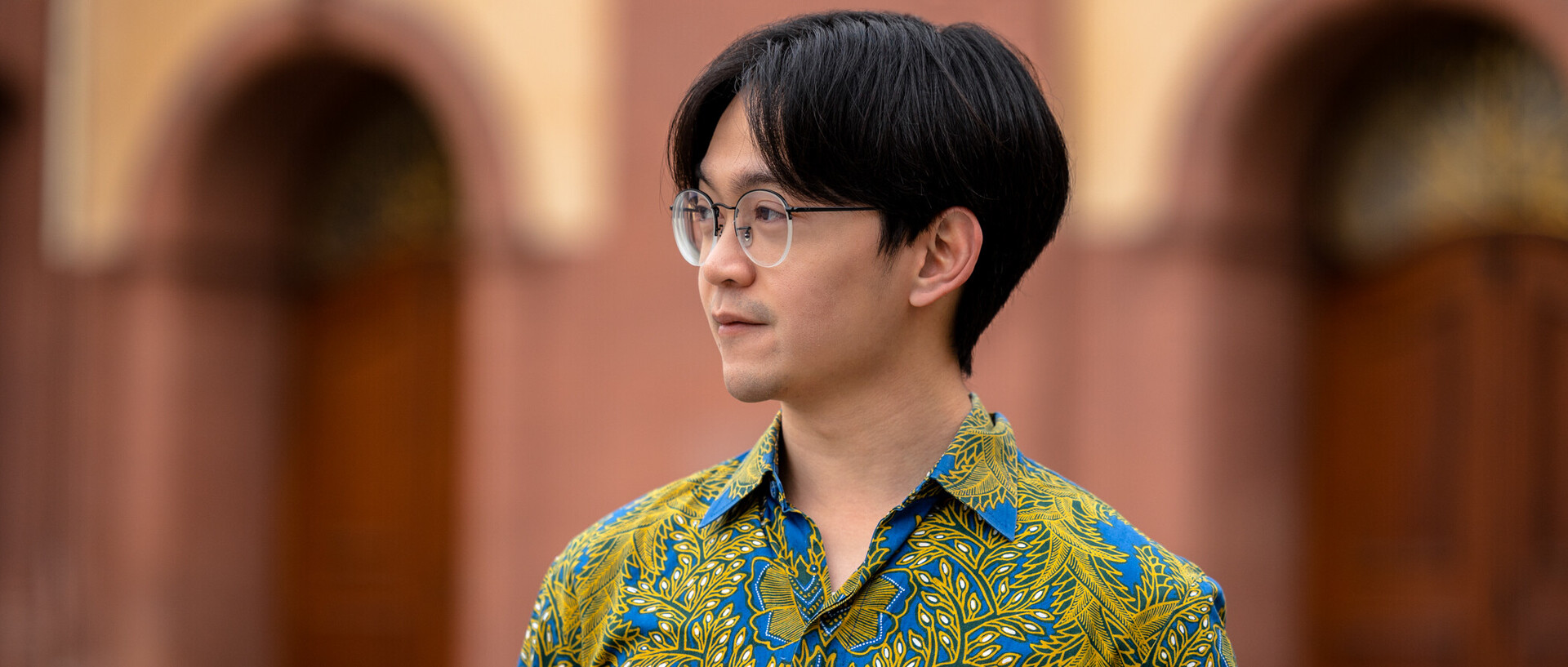 Ein Student mit Brille blickt zur Seite. Er trägt ein blau-grün gemustertes Hemd.