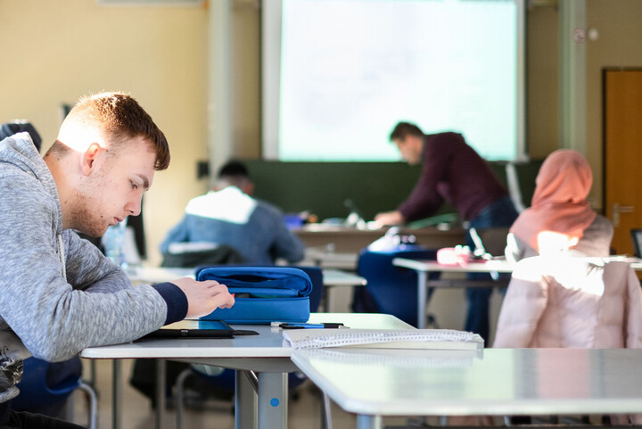 Students work with iPads in the classroom, with the teacher standing at the front.
