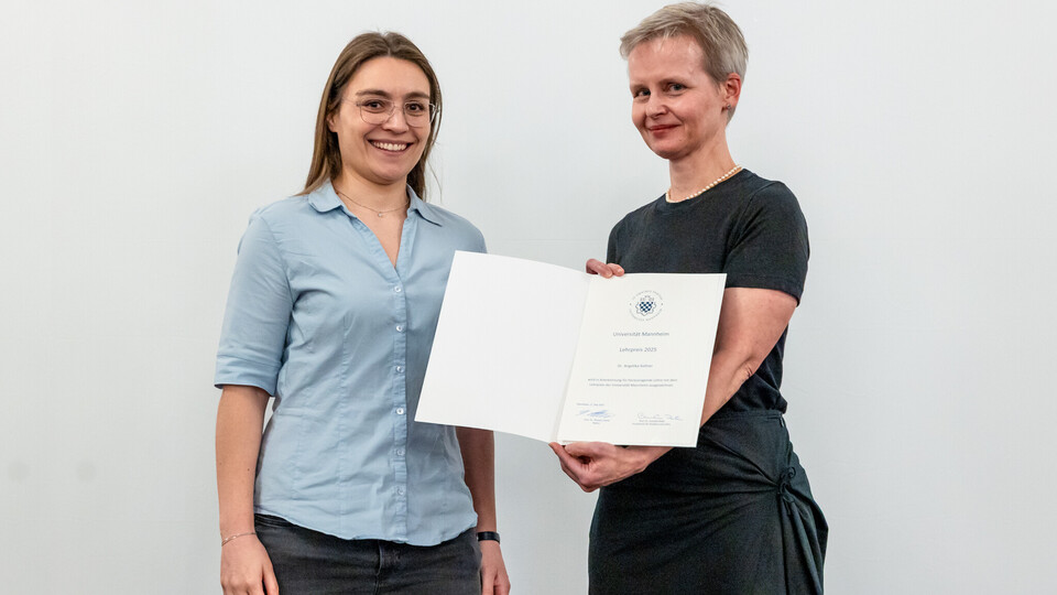Dr. Angelika Kellner, hellblaue Bluse, Brille und lange, braune Haare, erhält die Urkunde für den Lehrpreis von Prof. Dr. Cornelia Ruhe, die ein schwarzes Kleid trägt. Beide stehen nebeneinander und lächeln in die Kamera.