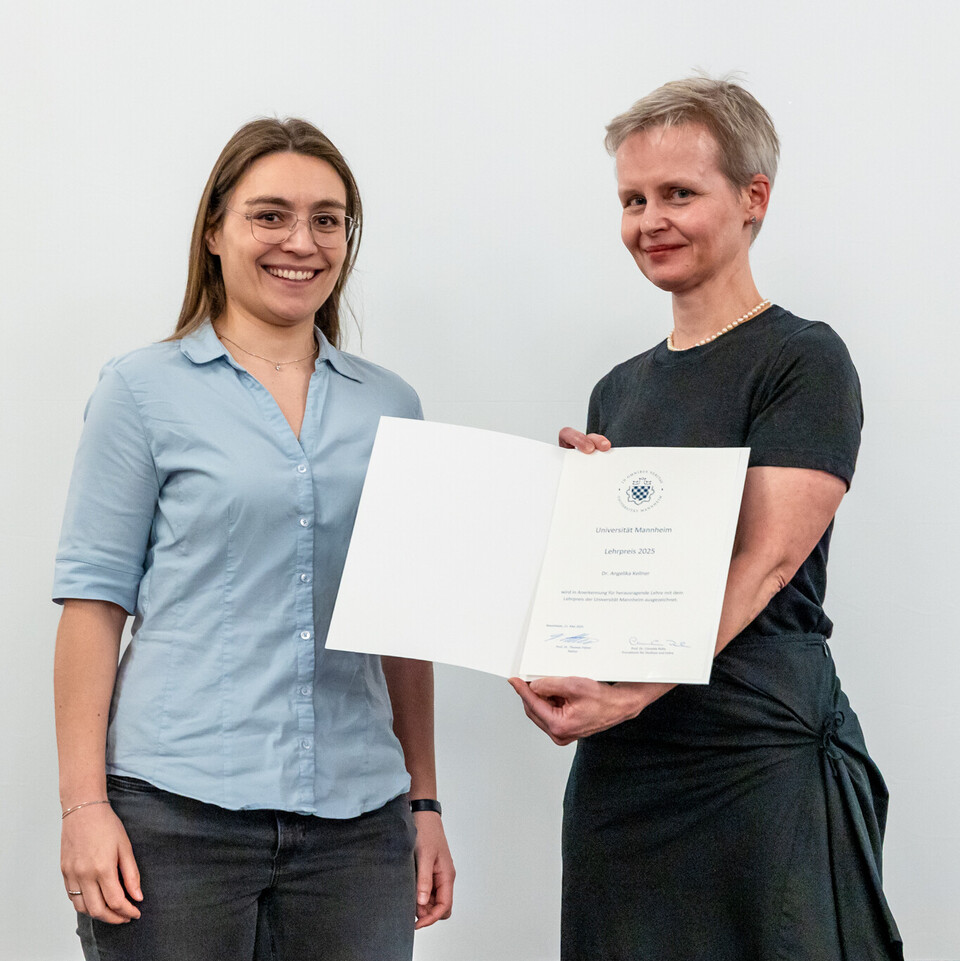 Dr. Angelika Kellner, hellblaue Bluse, Brille und lange, braune Haare, erhält die Urkunde für den Lehrpreis von Prof. Dr. Cornelia Ruhe, die ein schwarzes Kleid trägt. Beide stehen nebeneinander und lächeln in die Kamera.