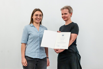 Dr. Angelika Kellner, hellblaue Bluse, Brille und lange, braune Haare, erhält die Urkunde für den Lehrpreis von Prof. Dr. Cornelia Ruhe, die ein schwarzes Kleid trägt. Beide stehen nebeneinander und lächeln in die Kamera.