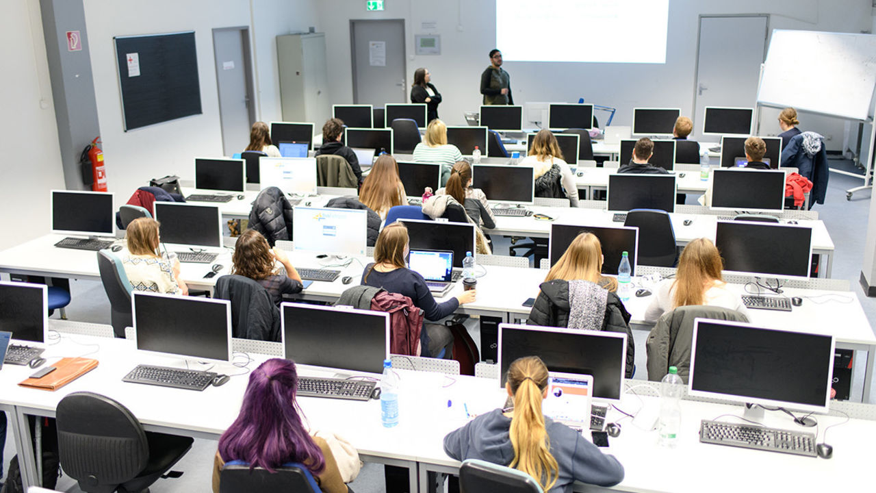 Studierende sitzen in einem hellen, weißen Computerraum. Ein Dozent hält einen Vortrag.