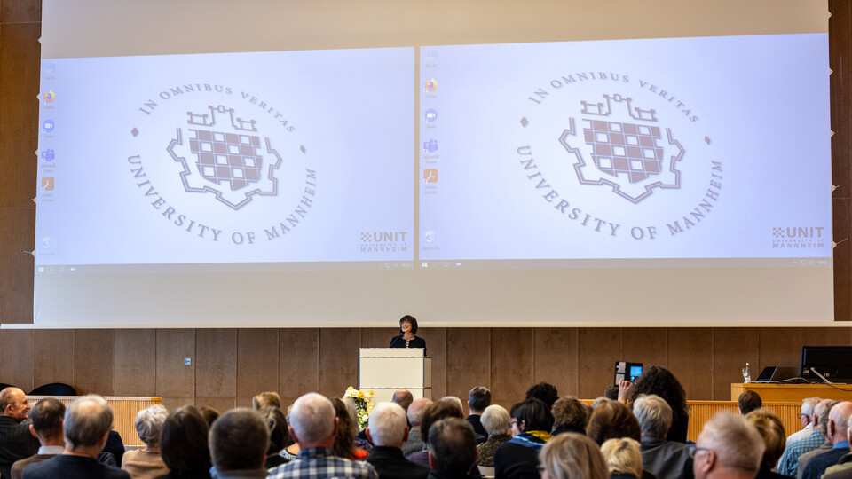 Foto von einer Veranstaltung des Gasthörendenstudiums in der Aula. Am Pult steht eine Referentin, vor ihr sitzen die Zuhörenden.