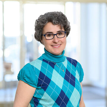 Susanne Quincke hat kurze graue lockige Haare. Sie trägt eine dunkelblau umrandete Brille und ein blaues kurzärmeliges Oberteil mit Argyle-Muster.