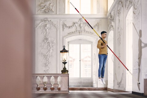 Student mit braunem Sweatshirt und blauer Jeans schwebt mit einem Fechtstab in der Hand in einem Festsaal des Schlosses.