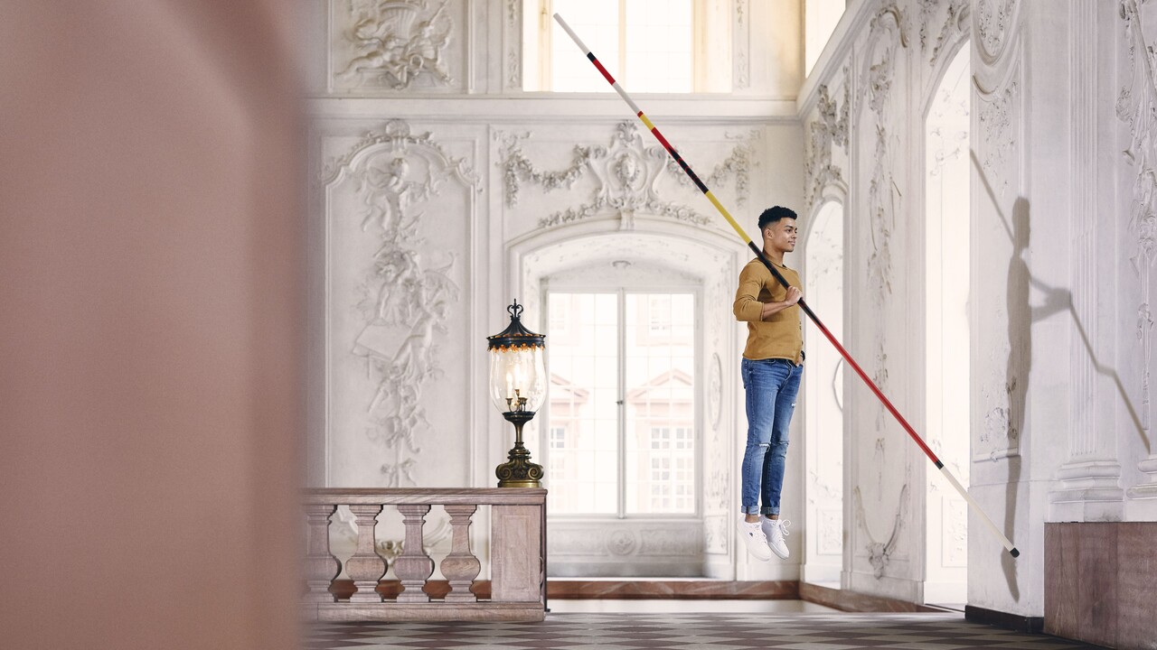 Student mit braunem Sweatshirt und blauer Jeans schwebt mit einem Fechtstab in der Hand in einem Festsaal des Schlosses.