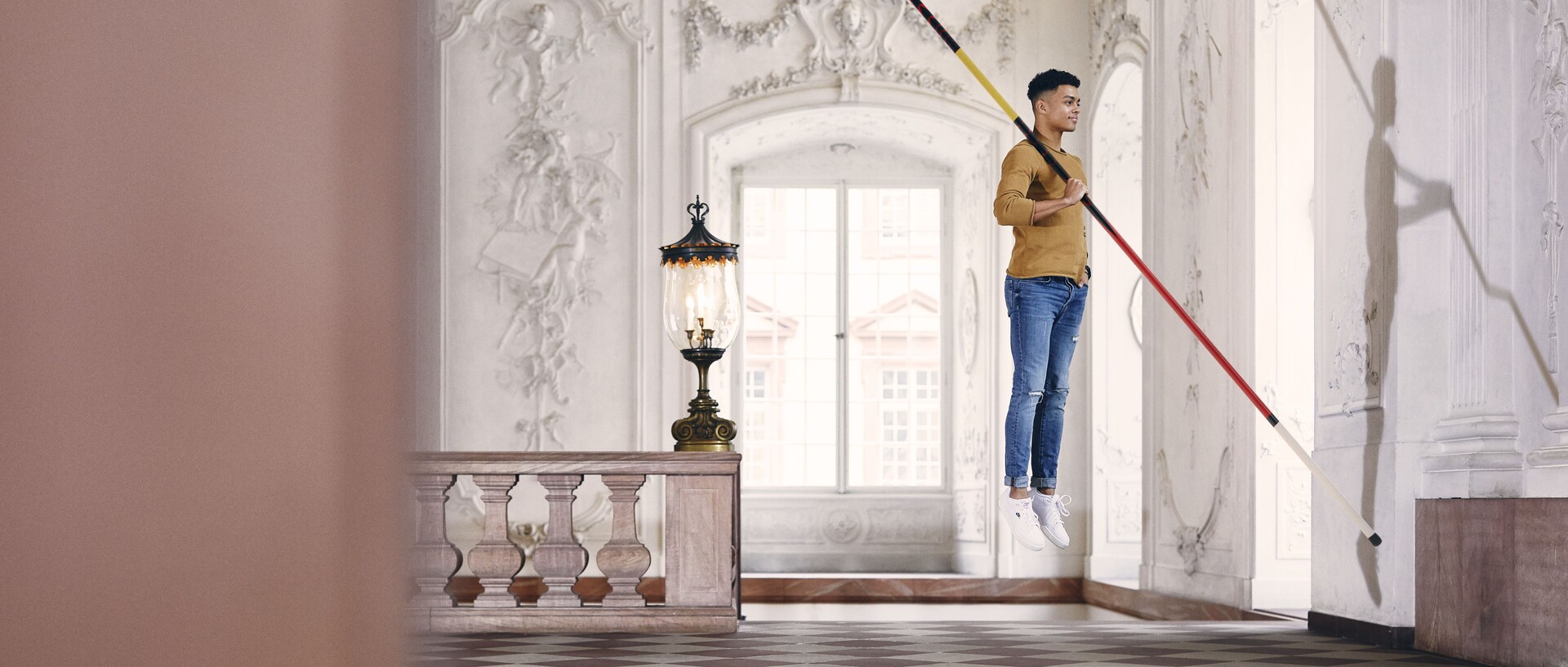 Student mit braunem Sweatshirt und blauer Jeans schwebt mit einem Fechtstab in der Hand in einem Festsaal des Schlosses.
