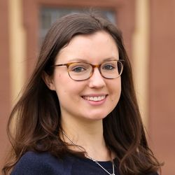 Melanie Waldmueller hat dunkelbraunes mittellanges Haar. Sie trägt einen dunkelblauen Strickpulli sowie eine Silberkette und eine Brille. Sie lächelt.