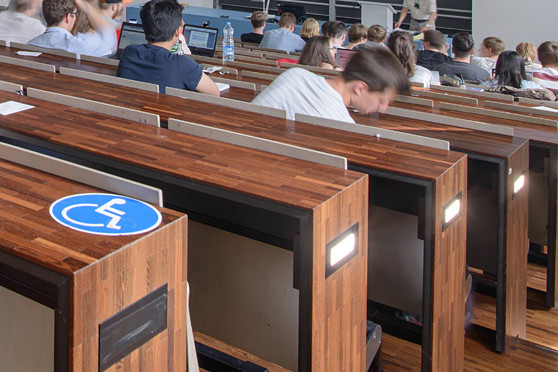 A lecture hall filled with students, an accesibility sign is glued to one of the tables 