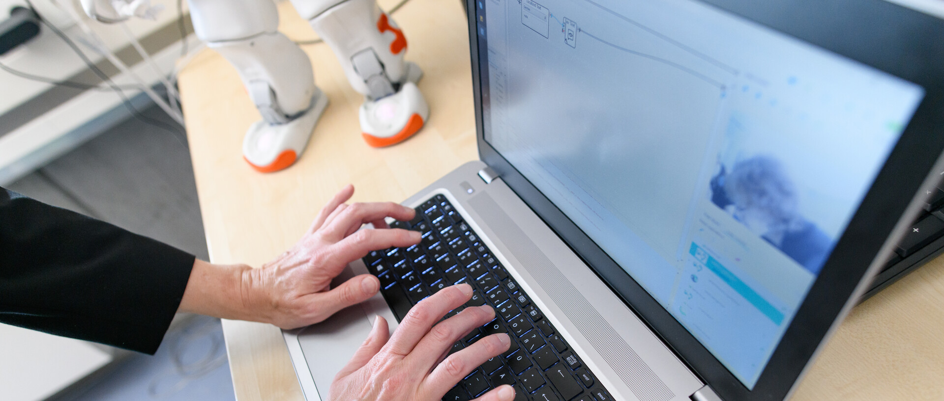 Eine Person arbeitet am Laptop. Im Hintergrund sind die Beine eines Roboters auf dem Schreibtisch zu erkennen.