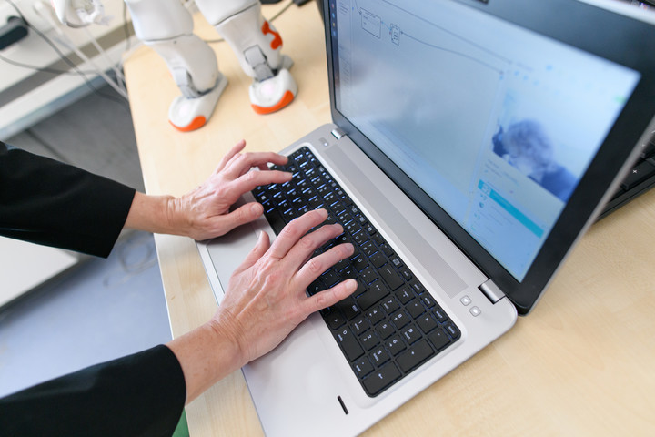 Eine Person arbeitet am Laptop. Im Hintergrund sind die Beine eines Roboters auf dem Schreibtisch zu erkennen.